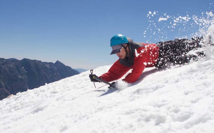 A person wearing a helmet appears to be sliding down a snowy incline on their belly. Snow is kicking up behind them.